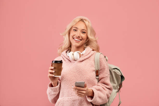 istock Pretty lady posing with backpack, holding coffee to go and cellphone, smiling at camera over pink studio background 1471885127
