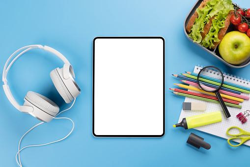 Tablet with blank screen, school supplies, stationery, and lunch box on blue background. Education and nutrition. Flat lay with blank space for your text or app