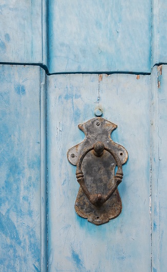 Antique rusty metal door knocker from the colonial period on an old wooden door with peeling paint.