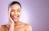Woman, smile and wink for skincare wellness or face dermatology beauty in purple studio background. Model, facial cosmetics care and natural glowing skin or comic happiness or headshot vision