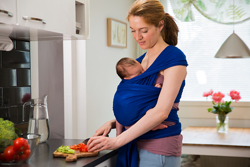 Woman with baby in a wrap carrier cooking in the kitchen, cutting vegetables with knife on a chopping board. Interior of a cozy modern kitchen. Mother doing her chores.