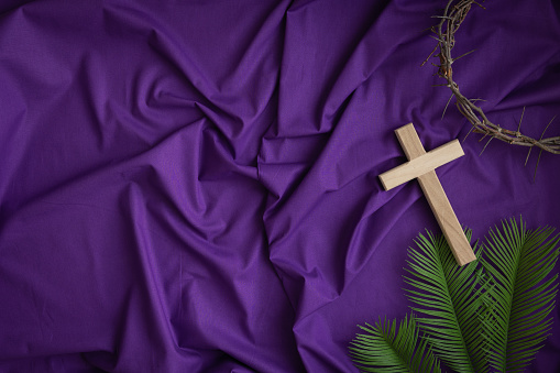 Border of simple wood Christian cross, partial crown of thorns and palm leaves on a dark purple fabric background with copy space
