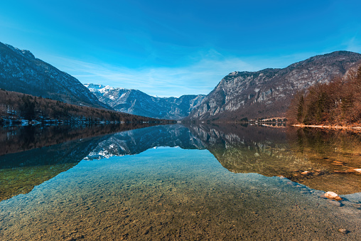 Beautiful scenic landscape of glacial Bohinj lake in Slovenia with crystal clear water and rocky shore on sunny winter morning with Julian Alps in background