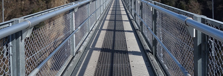 Pedestrian suspension bridge with steel grating and handrail.\nA suspension bridge is a type of bridge in which the deck is hung below suspension cables on vertical suspenders.\nAnti-slip steel grating (also known as slip-resistant grating) is a type of bar grating designed to prevent slippage. Steel bar grating is manufactured in a variety of methods. Welded bar grating consists of load bars electrically fuse welded to cross bars. Pressure-locked bar grating, or swaged grating, is where steel rods are forced through and locked in load bars. Riveted bar grating is where the load bars are riveted to the cross bars.