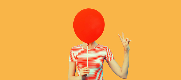 Portrait of funny woman covering her head with red balloon on orange background