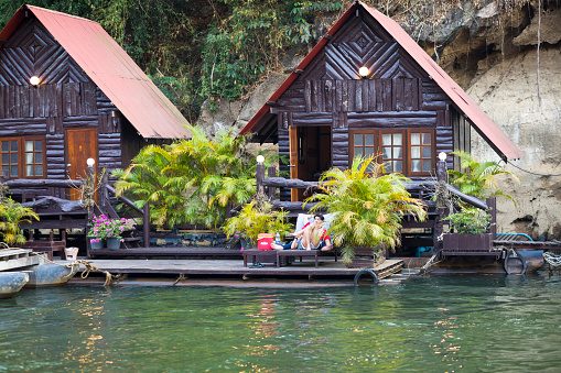 Relaxing Thai couple at wooden houses of a floating tourist resort on Kiwae Noi river in Kanchanaburi in Sai Yok National Park. Woman and man are relaxing on a sun lounger.  River is located in a canyon and offers idyllic water and nature vacations. In background is tropical rainforest