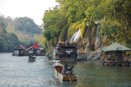 Queued boats and floating decks at waterfall of Kiwae Noi river  in Kanchanaburi in Sai Yok National Park. Most people are wearing life jackets. People have fun with water and are taking photos. In background is a floating tourist resort