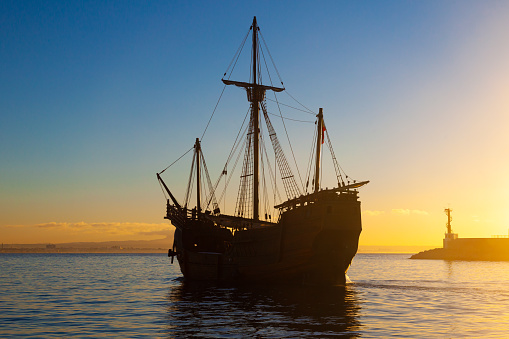 An old wooden caravel with folded sails leaving the sea bay at sunset at blue hour.