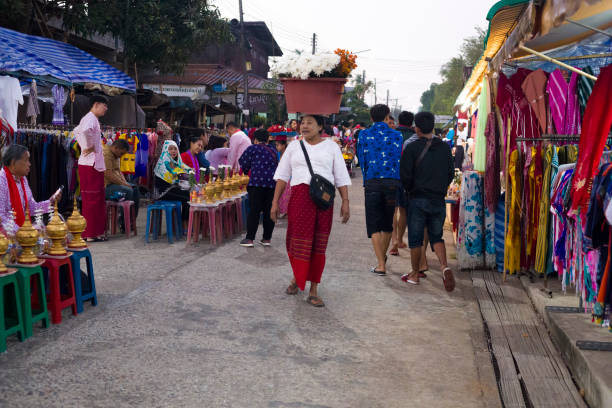 Thai Mon woman with flower basket on head is walking along  waiting Thai people with golden cauldrons at  ceremony of Makha Bucha Day in Sangkhla Buri Thai Mon woman with flower basket on head is walking along  waiting Thai people with golden cauldrons at  ceremony of Makha Bucha Day in Sangkhla Buri in northwest of Kanchanaburi province of Thailand in early morning close to sunset. People are wearing mostly traditional clothing and are standing in main street leading down to Mon Bridge and lake. People are preparing for monks coming along street true thailand classic stock pictures, royalty-free photos & images