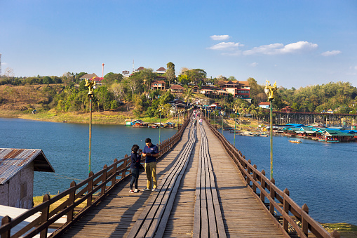 View along  historical Mon Bridge in Sangkhla Buri in west Thailand seated at lake and river near to border to Myanmar in Kanchanaburi province and northern Khao Laem national Park. People are on bridge in foreground and background. In background are buildings on hills