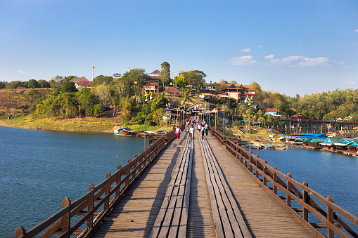 Thai people are walking  along  historical Mon Bridge in Sangkhla Buri in west Thailand seated at lake and river near to border to Myanmar in Kanchanaburi province and northern Khao Laem national Park. People are on bridge in  background. In background are buildings on hills