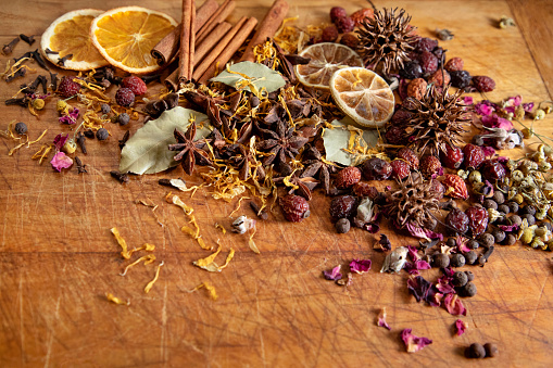 A variety of spices and petals including cloves, star anise, bay leaves, cinnamon, allspice, rose petals, calendula petals, rose hips, dried lemon slices, and dried orange slices thrown on a rustic cutting board.