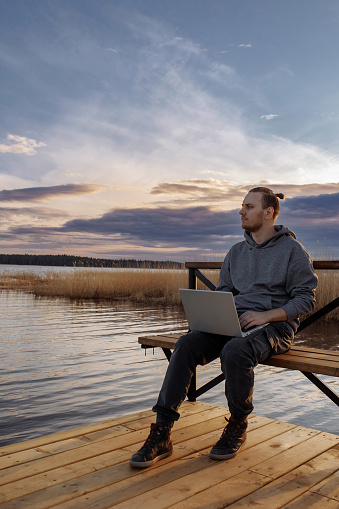A handsome young man is resting and working on a laptop near the sea. A freelancer works sitting on a wooden bench on a pier by the water. Looks into the distance.