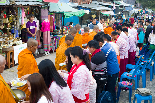 Traditionally dressed thai people are giving food to monks   at  ceremony of Makha Bucha Day in Sangkhla Buri in northwest of Kanchanaburi province of Thailand in early morning close to sunset. People are wearing mostly traditional clothing and are standing in main street leading down to Mon Bridge and lake. People are waiting to give food donations to monks when they pass along street down to bridge