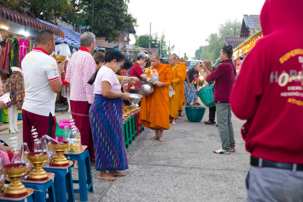 Adult Thai men and women  are giving food donations to  thai monks at  ceremony of Makha Bucha Day in Sangkhla Buri Adult Thai men and women  are giving food donations to  thai monks at  ceremony of Makha Bucha Day in Sangkhla Buri in northwest of Kanchanaburi province of Thailand in early morning close to sunset. People are wearing mostly traditional clothing and are standing in main street leading down to Mon Bridge and lake. People are waiting to give food donations to monks when they pass along street down to bridge true thailand classic stock pictures, royalty-free photos & images