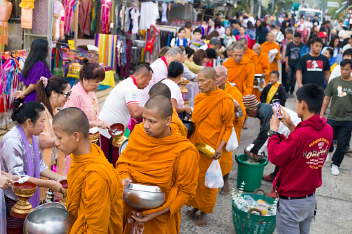 View over heads of rows of thai people and monks  giving  food donation to thai monks  at  ceremony of Makha Bucha Day in Sangkhla Buri in northwest of Kanchanaburi province of Thailand in early morning close to sunset. People are wearing mostly traditional clothing and are standing in main street leading down to Mon Bridge and lake. People are waiting to give food donations to monks when they pass along street down to bridge