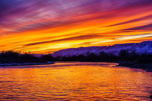 Dramatic Sunset Reflections in Water with Mountain backdrop - Snow-covered mountains of the Roan Plateau with dramatic vibrant sunset colors reflecting in the Colorado River. Silt, Colorado USA.