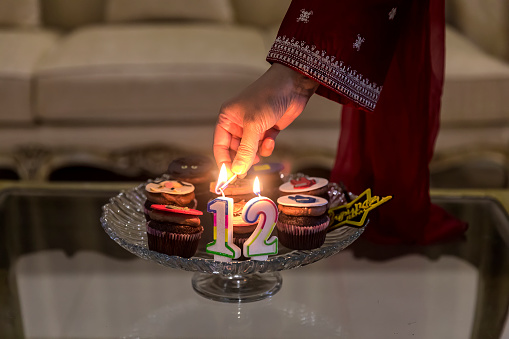 The photo depicts a woman lights up the candles for the 12th anniversary celebrations while the  candles flicker and dance in the dim light, casting a warm glow over the room. The scent of freshly baked cupcakes and muffins fills the air, making your mouth water in anticipation. The cakes are decorated with colorful frosting and sprinkles, making them look almost too pretty to eat.