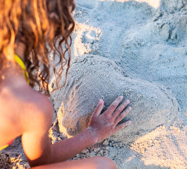 Young girl lies on the beach, covered in sand while pretending to be a mermaid under the sun.