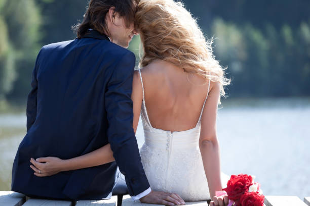 couple of young women wearing their wedding dresses while sitting with their backs to each other on a pier at the seashore