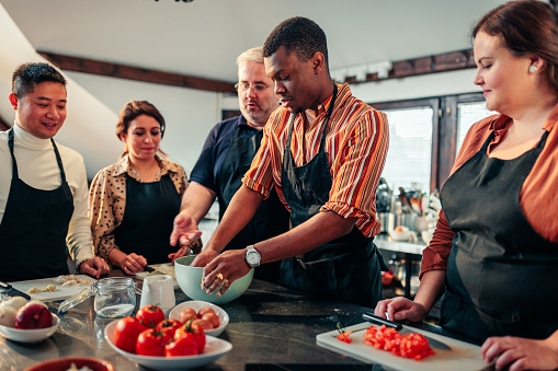 A young African American man is in a group cooking class, making a meal, with the other students and the teacher watching him.