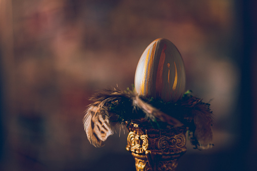 Dark low key still life with directed light from the left of a golden and pastel green painted Easter egg on green moss and brown feathers on top of an ancient colored pillar with copy space. Creative color editing with added grain. Very soft and selective focus. Part of a series.