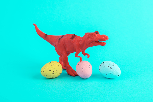 Toy red dinosaur tyrannosaurus rex with easter colored eggs on a turquoise background. Minimalism creative layout