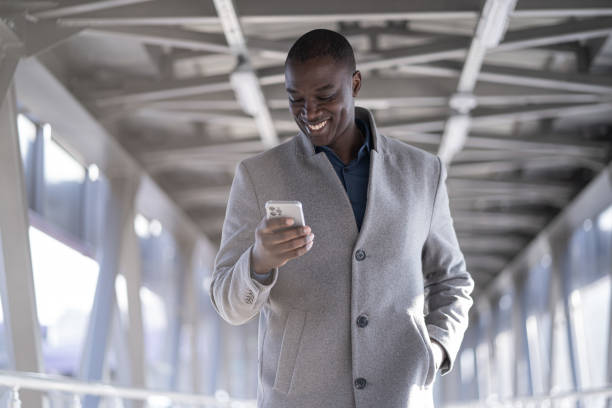 feliz africano - hombre americano mirando su teléfono inteligente, mientras camina por un paso elevado. joven parado al aire libre sobre una estructura metálica. - businessman 30s low key surprise fotografías e imágenes de stock