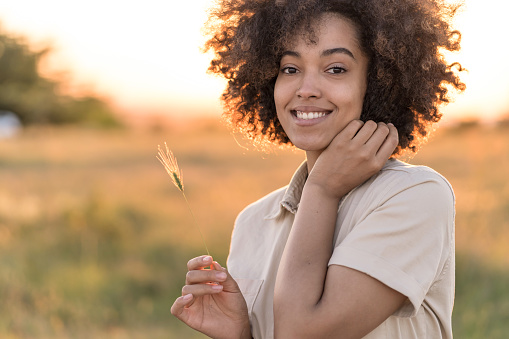 Portrait of beautiful african american woman holding a grass straw, smiling and looking at the camera in front of grass field during sunset. Outdoor portrait of a smiling multiracial girl. Happy cheerful girl laughing in the nature