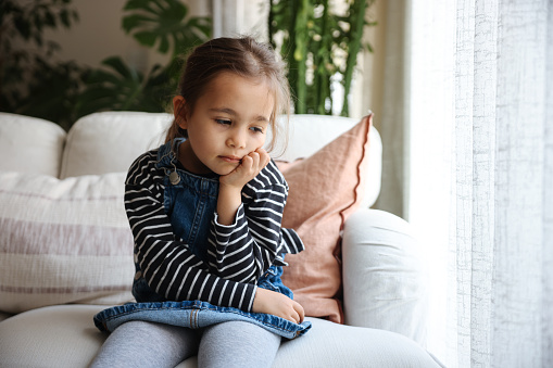 Close up portrait of pensive serious little school preschool age girl standing by window at home looking at camera with sad unhappy face having problem trouble thinking about bad relations in family