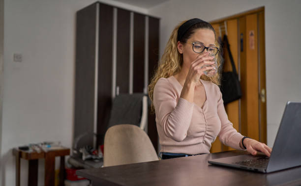 istock Young woman working from home, looking at laptop screen while drinking water 1471945899