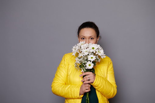 Portrait of a charming dark-haired woman with a bouquet of white spring flowers, looking confidently at camera, isolated over gray background. Beautiful women. People. Lifestyle. Festive event.