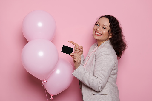 Hispanic woman in elegant light gray jacket, holds a credit card, standing on isolated pink background with inflatable pastel balloons, expresses positive emotions, smiling with beautiful toothy smile