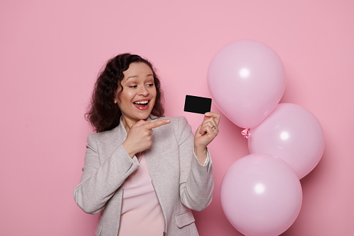 Smiling African American middle-aged woman in light gray formal suit, pointing at a black credit card, isolated on pink background with inflatable pink helium balloons. Banking Business People concept