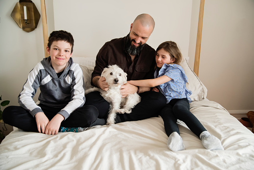 Father and kids portrait on a bed at home. They are casually dressed for the week-end, father has a beard and a bald head, kids are 8 and 10 year’s old. Dog is a mix of a west highland terrier and a maltese. Horizontal full length shot with copy space. This was taken in Montreal, Quebec, Canada.