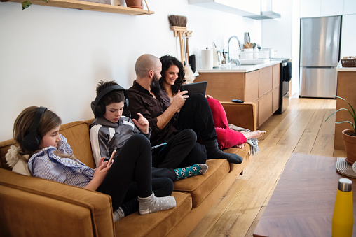 Kids are playing video games, step-mom and dad are shopping on mobile devices on the living room couch. They are casually dressed for a week-end at home. Horizontal full length indoors shot with copy space. This was taken in Montreal, Quebec, Canada.