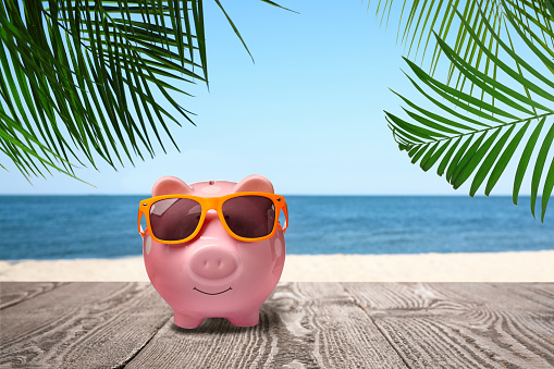 Saving money for summer vacation. Piggy bank with sunglasses on wooden surface near sandy beach and sea