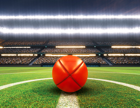A red aussie rules ball on the center line of a stadium with posts on a marked green grass pitch at night under illuminated floodlights - 3D render
