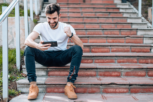 A young Caucasian man is sitting on a flight of stairs and cheerfully watching a sports stream on his phone.