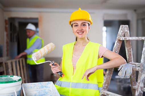 Portrait of smiling woman painter wearing hardhat and yellow vest. Renovation works in house.