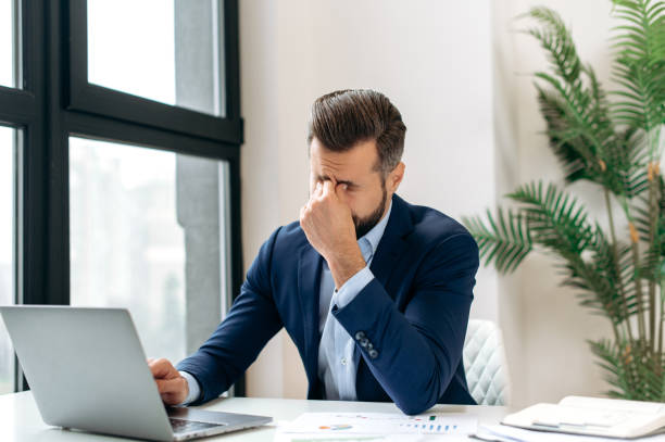 istock Overworking. Frustrated tired caucasian  entrepreneur, product manager, programmer, sit at a desk in the office, massaging the bridge of his nose with his eyes closed, feeling exhausted from long work 1471974241
