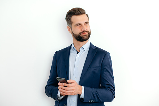 Successful caucasian business man in elegant clothes, stands on a white isolated background, uses his smartphone, chats online on social networks, looks to the side, thinking, planning