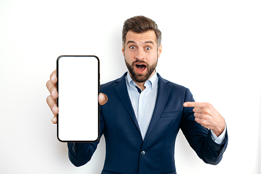 Shocked amazed surprised caucasian business man, in a suit, showing smart phone with empty mock-up screen in hand for advertising, points finger at it, standing on isolated white background