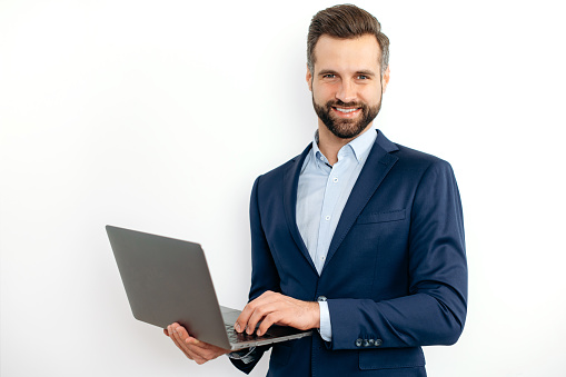 Friendly attractive caucasian bearded business man, in a suit, programmer, IT specialist, holding opening laptop in hands, standing on isolated white background, looks at camera, smiling