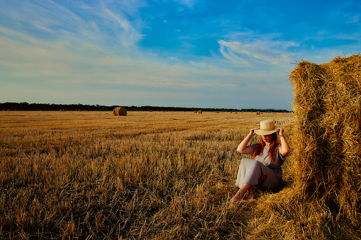 A beautiful young woman in a hat and a summer dress is sitting on a sheaf of hay in a field. Rural nature, wheat field, rest in the country, unity with nature