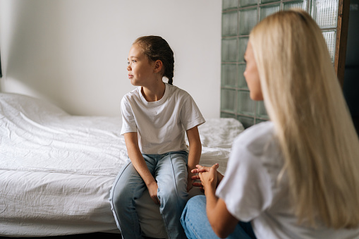 Back view from unrecognizable young mother talking counseling upset offended pretty child girl feels sad insulted sitting on bed at home. Concept of mom-children problem in preteen years.