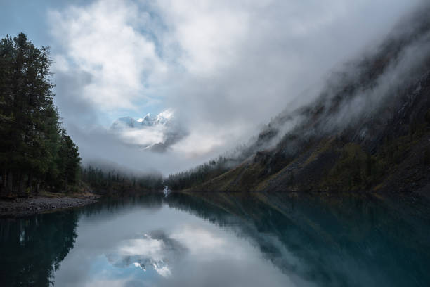 istock Tranquil scenery with snow castle in clouds. Mountain creek flows from forest hills into glacial lake. Snowy mountains in fog clearance, small river and coniferous trees reflected in calm alpine lake. 1471978948
