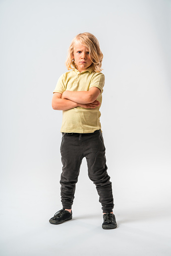 A blonde boy with long hair, dressed in light brown shirt and black pants. Posing in front of a white studio background and looking angrily at the camera, his arms crossed. Full length image.