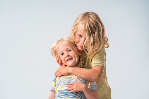 Two blond, long-haired brothers holding each other with love and posing for the camera. Waist up shot in front of a white studio background. Looking at the camera.