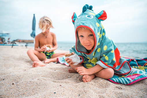 Young caucasian siblings sitting on sand, eating sandwiches and wearing swimwear. The youngest kid is looking at the camera, wearing a dragon hooded towel.